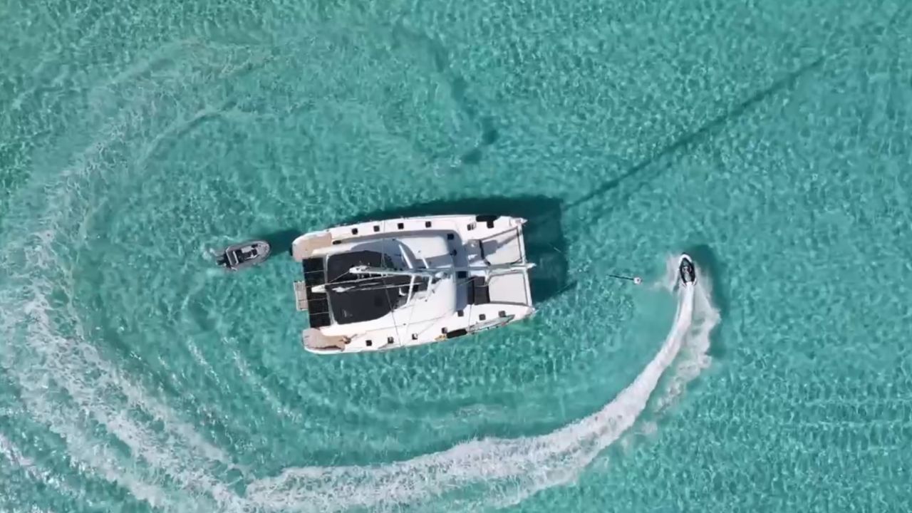 Aerial view of a sailing catamaran in pale turquoise water with a curving wake from a personal watercraft