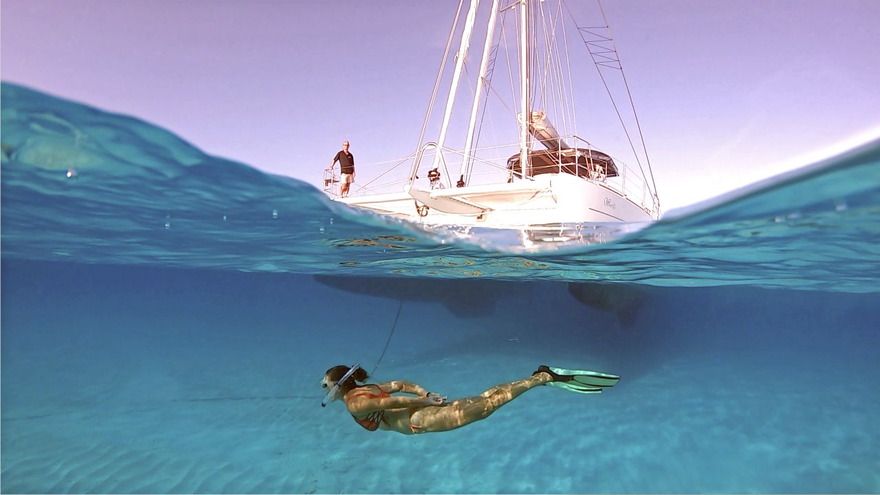 Split-level view of a sailing catamaran at anchor above clear water with a snorkeler gliding beneath