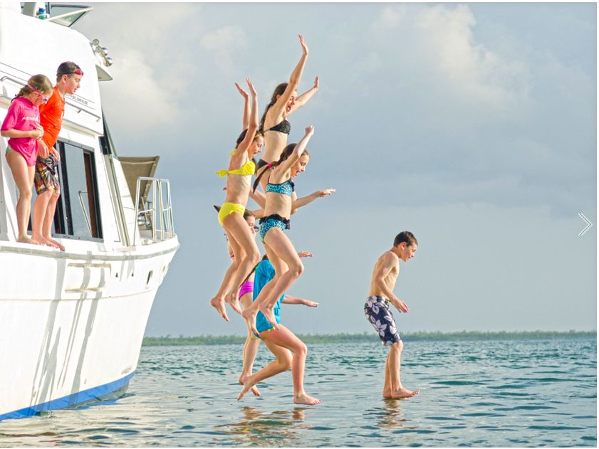 Children jumping from a boat into shallow, clear water as others watch from the deck